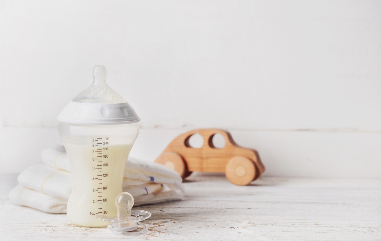 baby bottle of goat milk formula placed on a counter top with a wooden baby toy and baby blankets